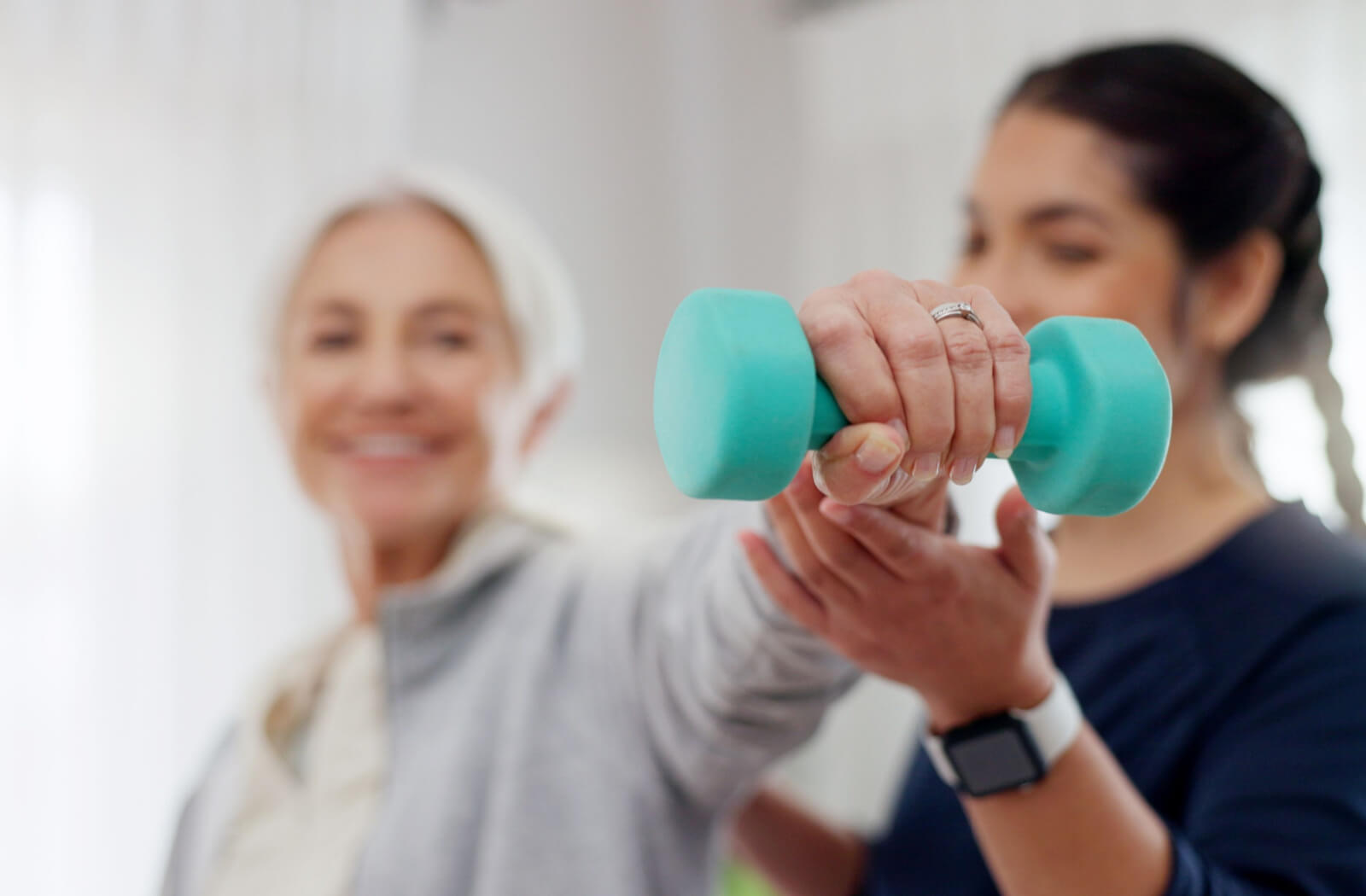 A professional caregiver supports an older adult as they lift a small blue handheld weight to help with their arthritis
