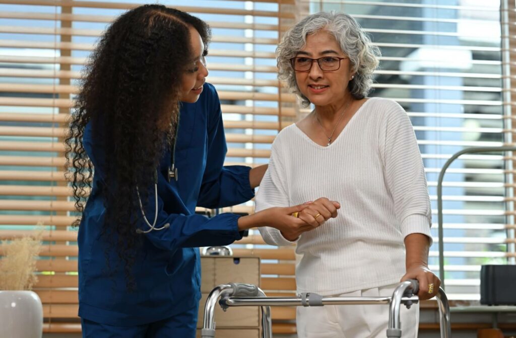 A nurse holds an older adult's hand while helping them move around with the aid of a walker while recovering from a stroke