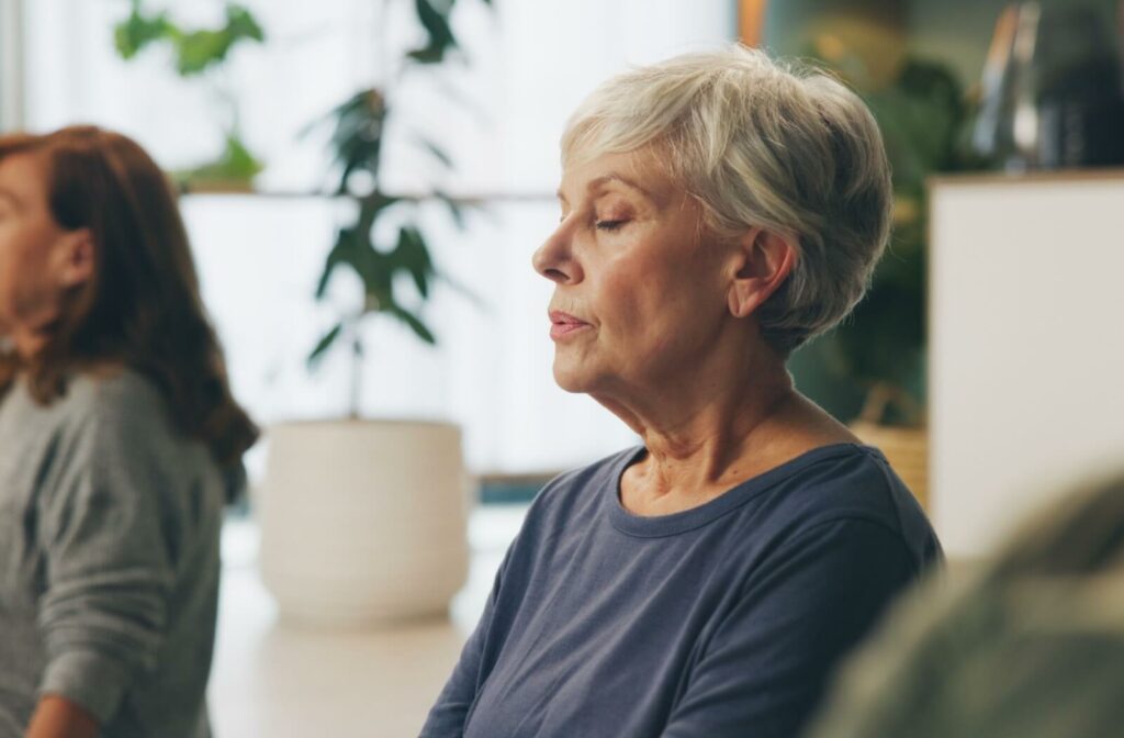 An older adult closes their eyes and breathes slowly out of their mouth during an indoor group meditation to support mental health